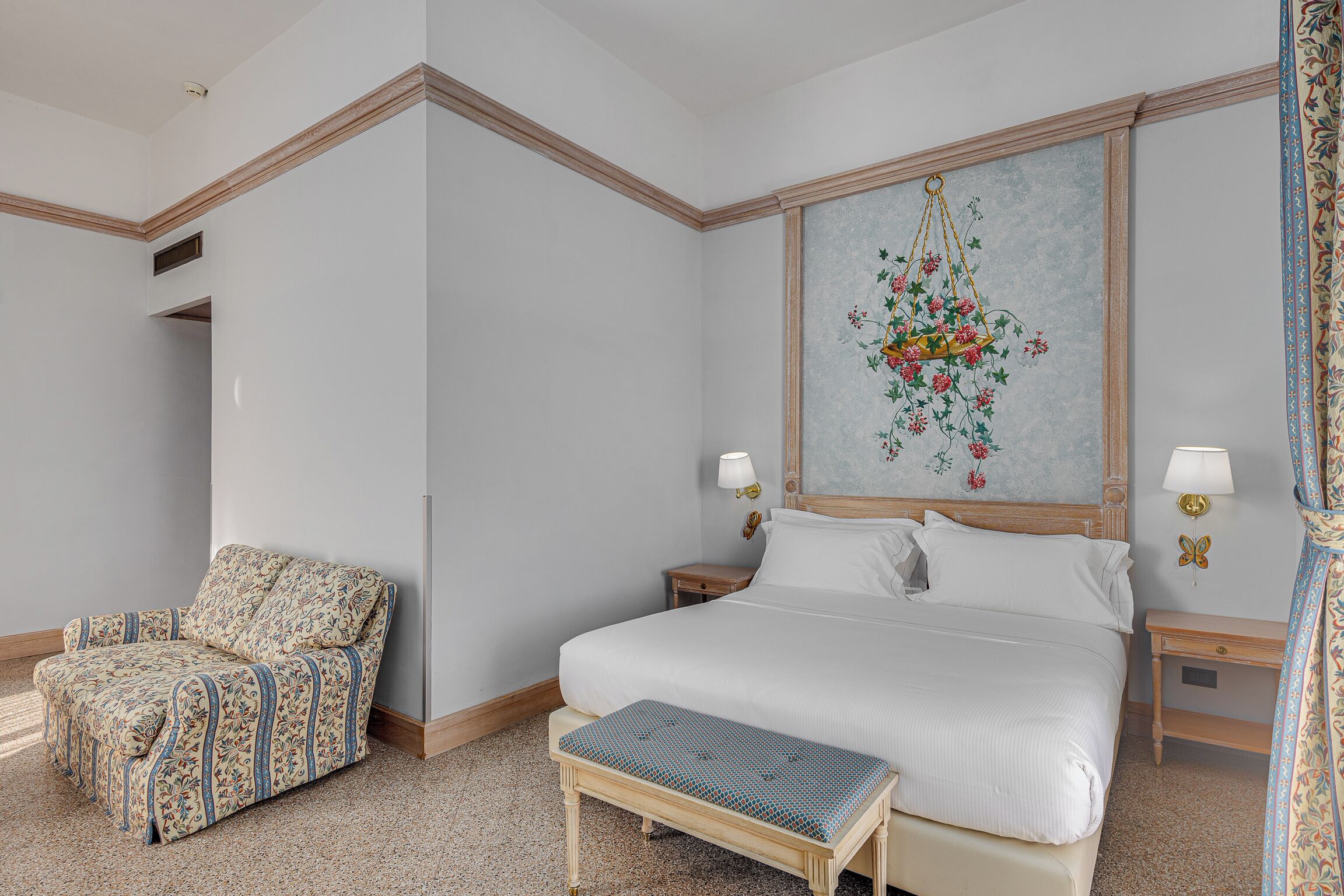 Quarto de hotel com uma cama grande, uma cadeira, um banco e uma parede com um quadro floral.