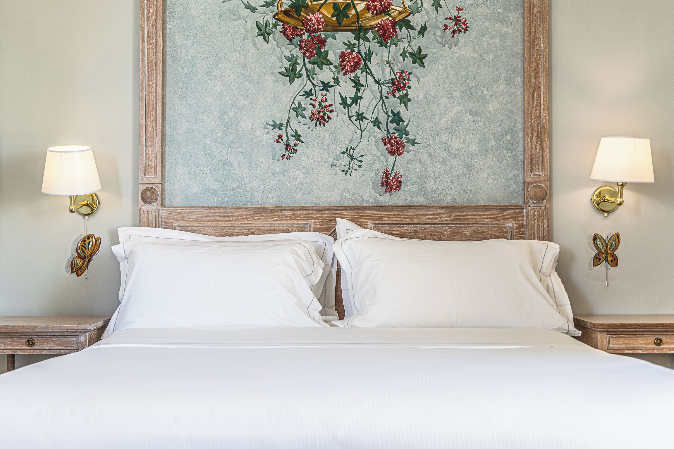 Een net opgemaakt bed met witte lakens, een houten hoofdbord en een bloemschilderij erboven. Aan weerszijden van het bed hangen twee wandlampen met vlinderdecoraties.