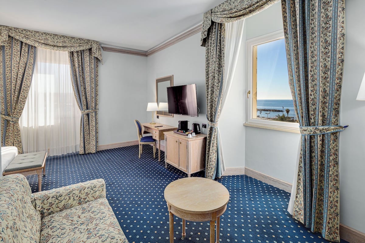 Een hotelkamer met een bed, een bureau met een televisie, een kleine ronde tafel en uitzicht op zee door een raam.