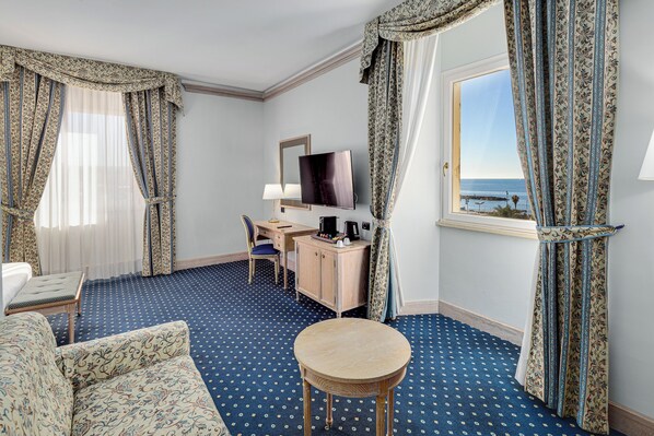 Habitación de hotel con cama, escritorio con televisor, una mesita redonda y vista al mar a través de una ventana.