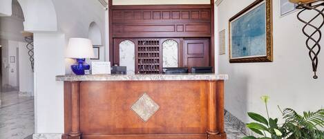 A reception area with a marble floor, a wooden reception desk, a blue lamp, and a framed painting on the wall.