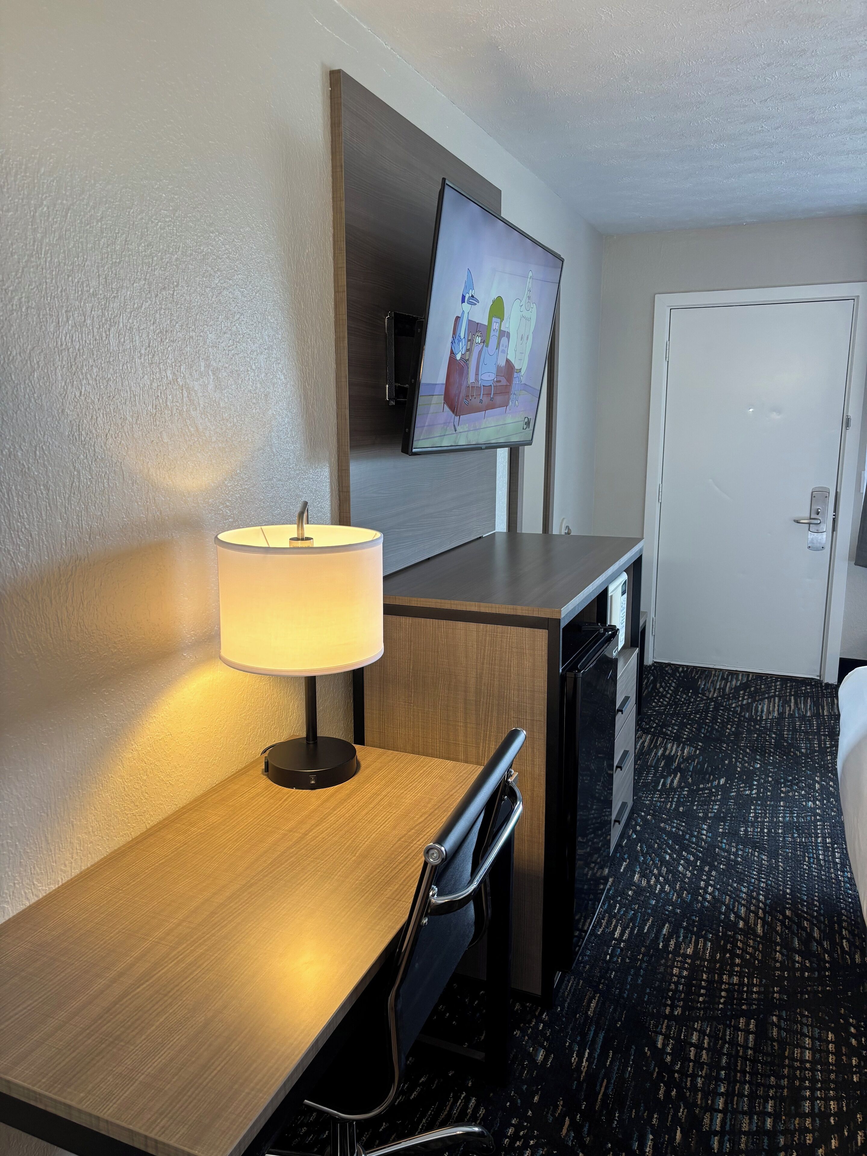 A hotel room with a wooden desk, a lamp, a television mounted on the wall, a white door, and a patterned carpet.
