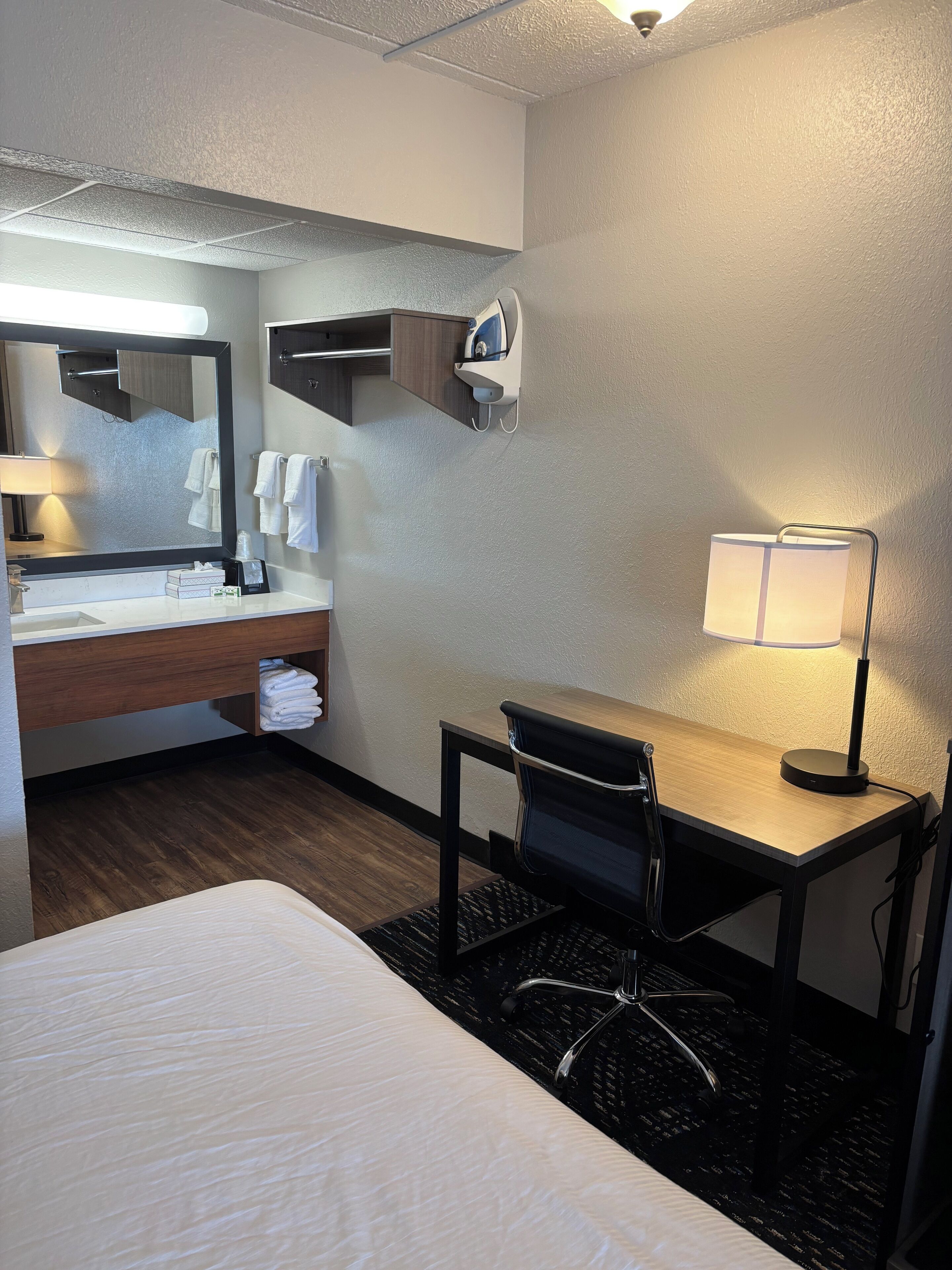 A hotel room with a bed, a desk with a chair, a desk lamp, a mirror, a towel rack, and a wall-mounted hairdryer.
