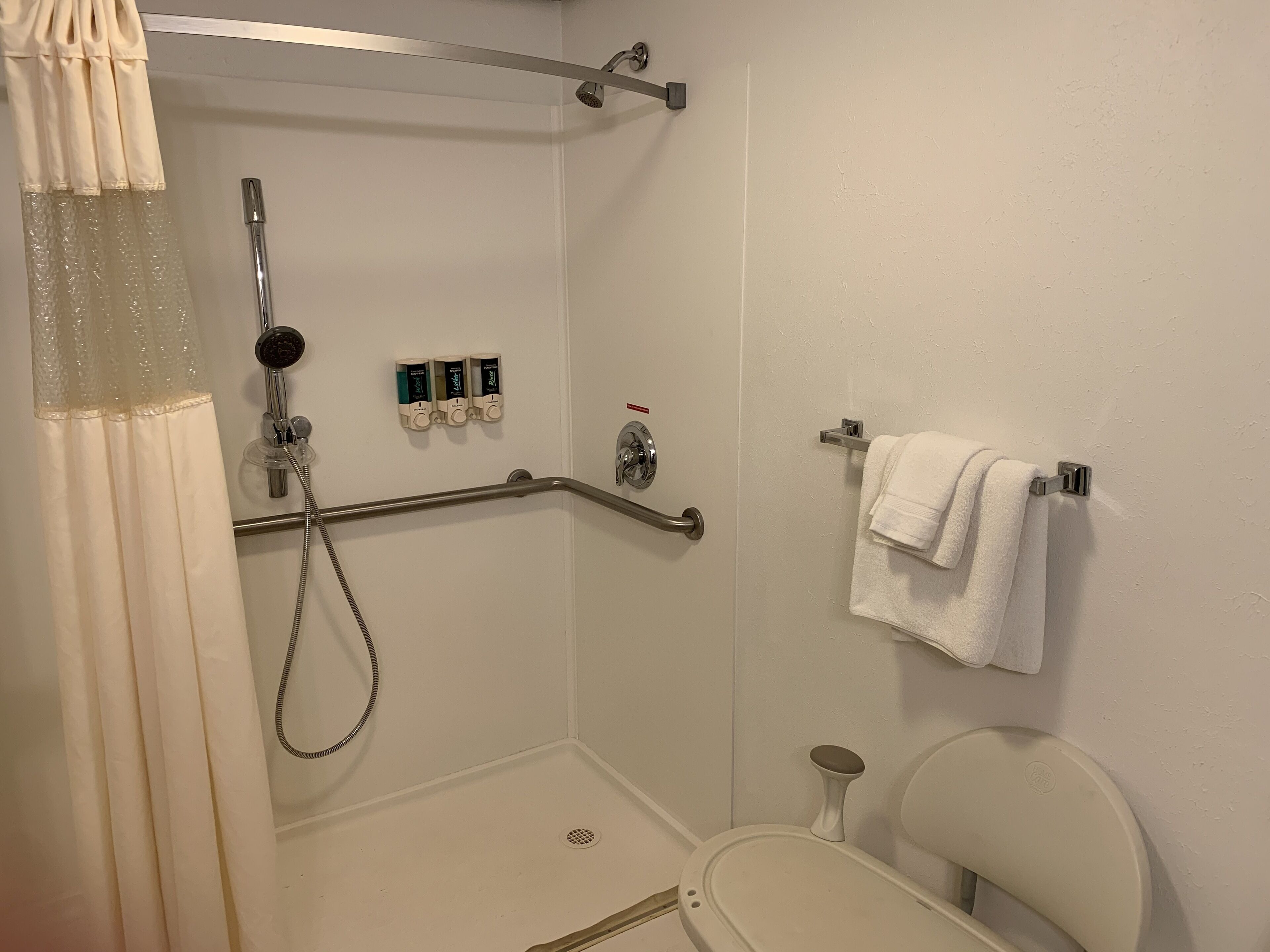 Um chuveiro acessível para cadeirantes, com barra de apoio, vaso sanitário branco e porta-toalhas com toalha branca.