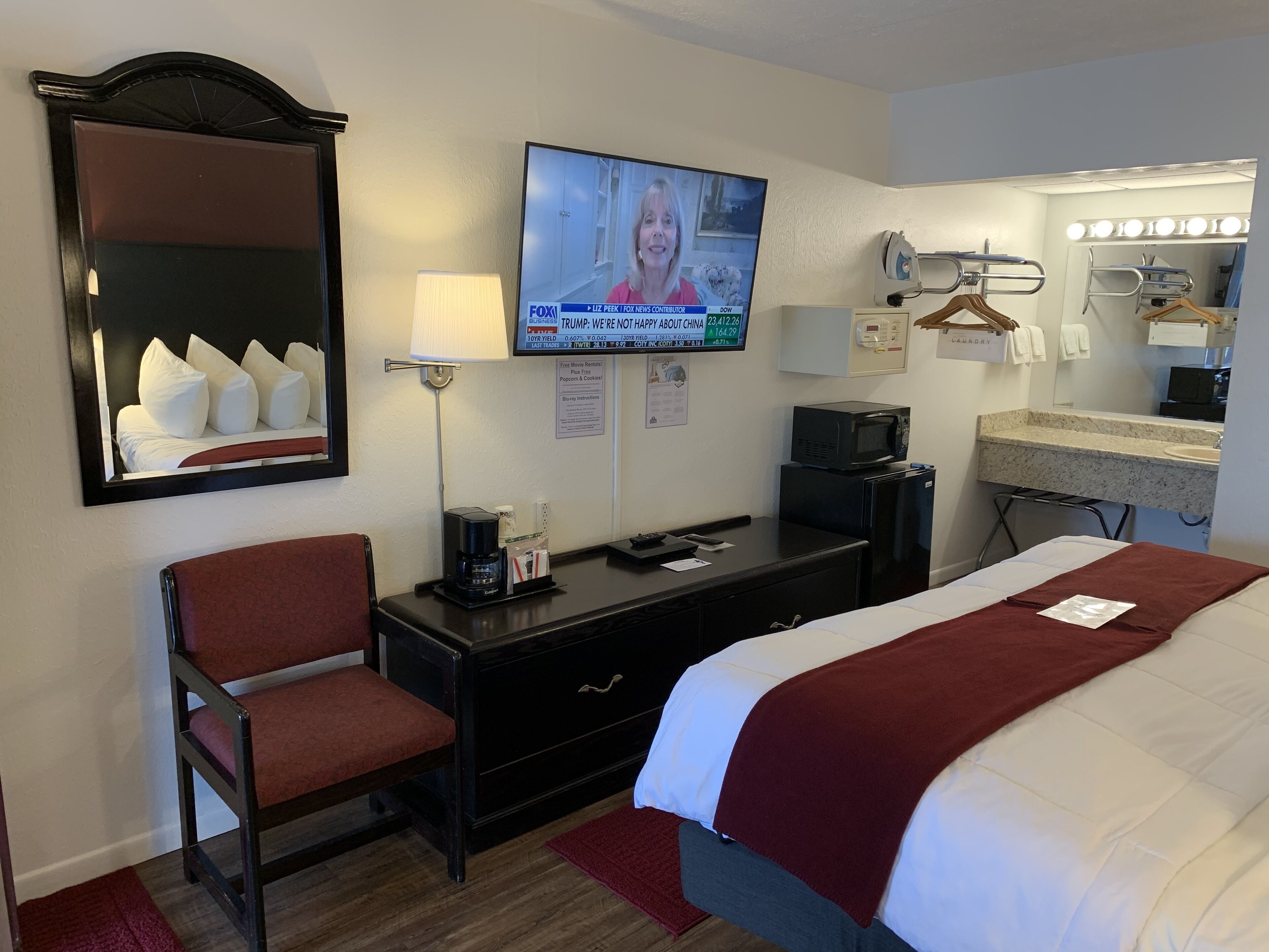 Una habitación de hotel con cama, escritorio, silla, televisor, espejo, lámpara, microondas y refrigerador pequeño.