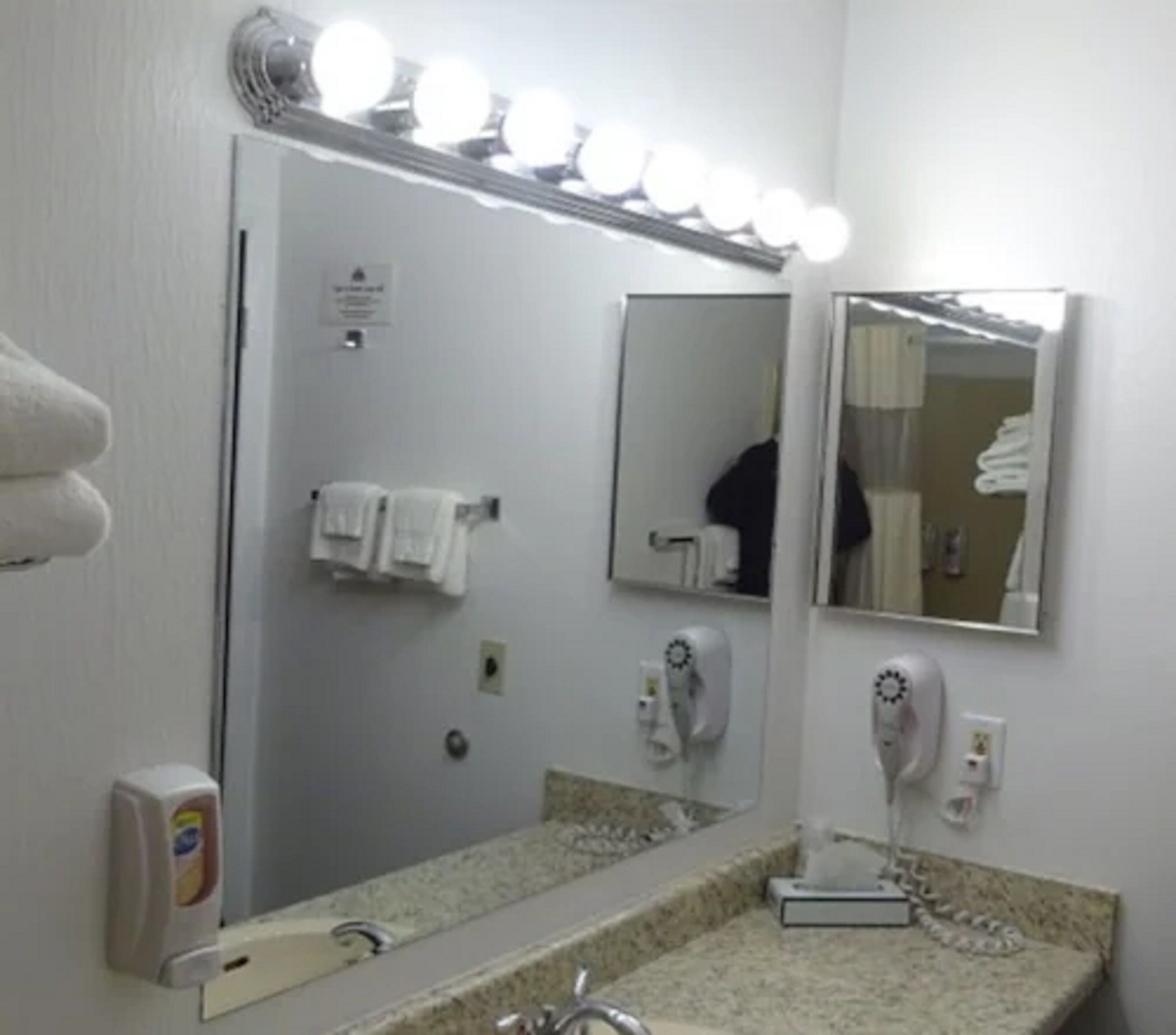 Une salle de bain avec un grand miroir, des luminaires au-dessus, un sèche-cheveux et un porte-serviettes.