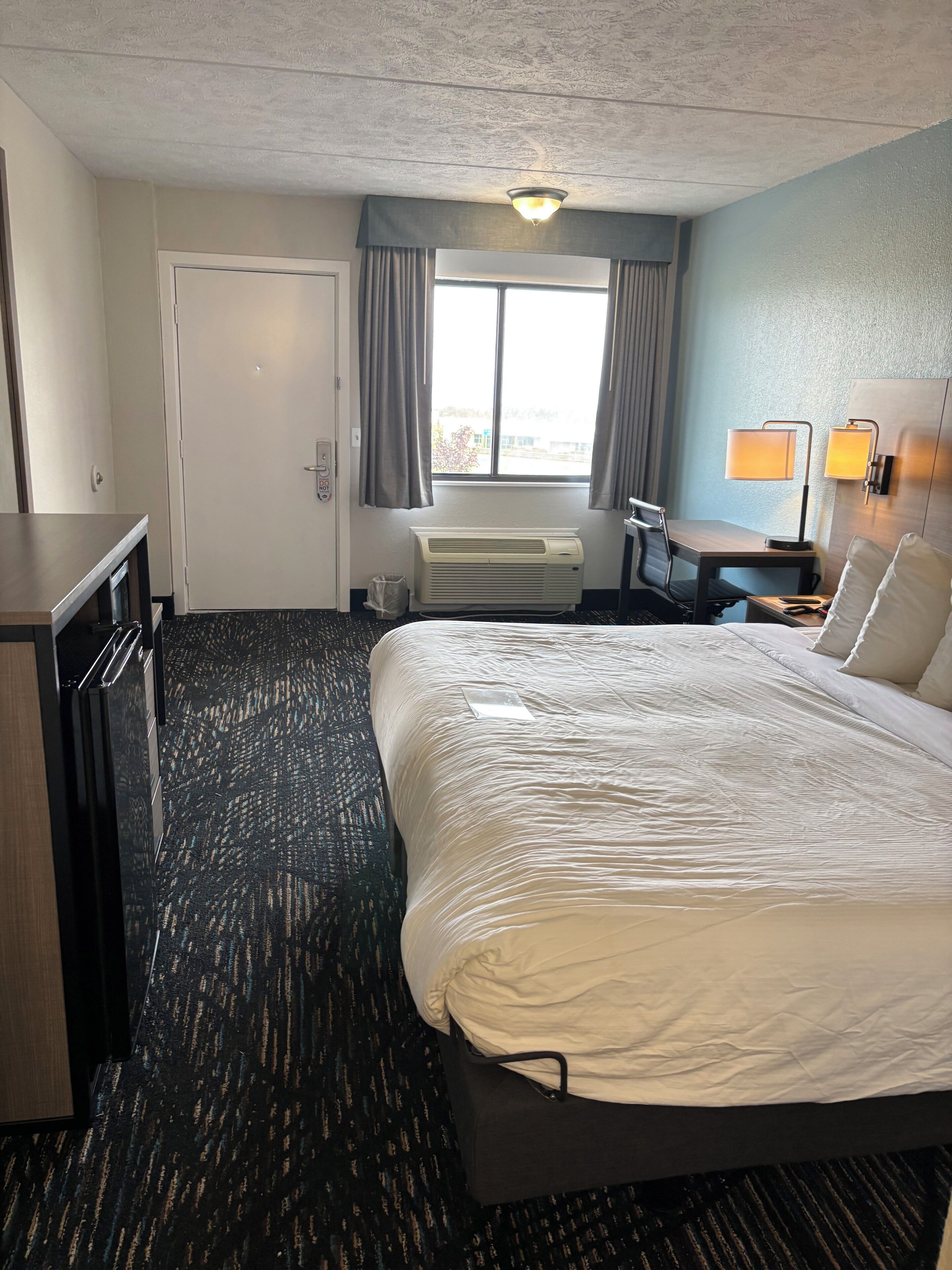 A hotel room with a large bed, a desk, a chair, a window with curtains, a wall-mounted lamp, and an air conditioning unit.
