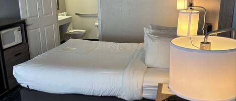 A hotel room with a bed, bedside table, lamp, and a view of the bathroom.