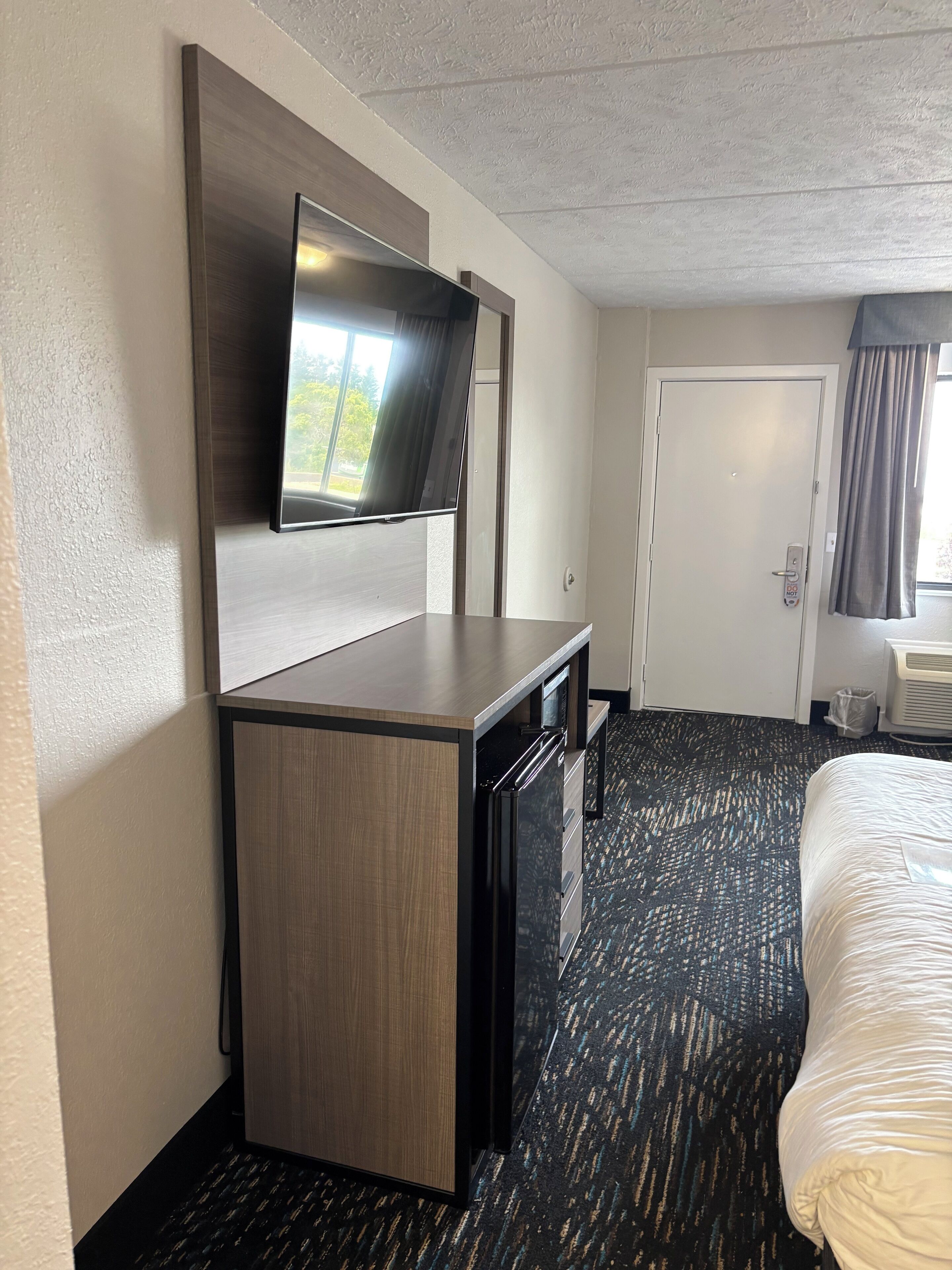 Habitación de hotel con televisión de pantalla plana montada en un mueble de madera, cama con colcha blanca y suelo alfombrado.