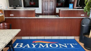 Ein Empfangsbereich mit Baymont by Wyndham Logo auf einer blauen Matte, einem Holzbüro, zwei Monitoren, einem Mülleimer und einem Blumentopf.