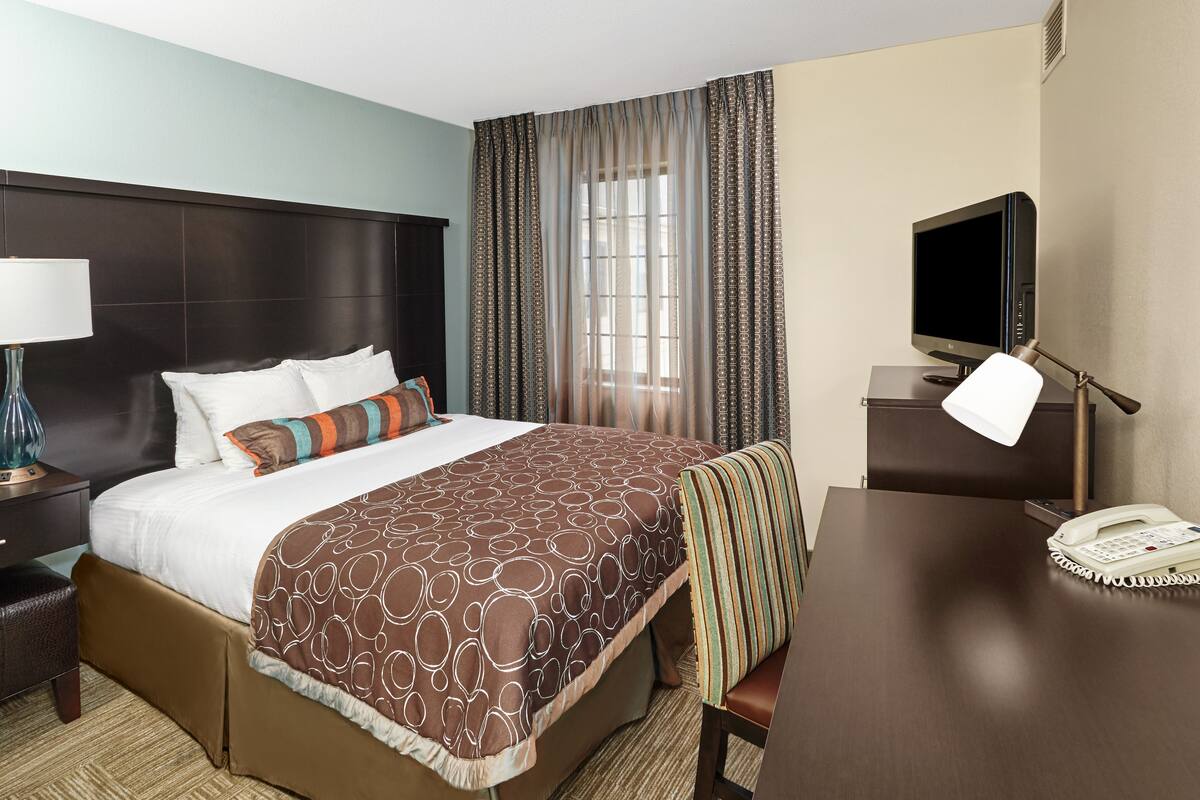 A hotel room with a large bed, a desk with a telephone, a television, and a nightstand with a lamp.