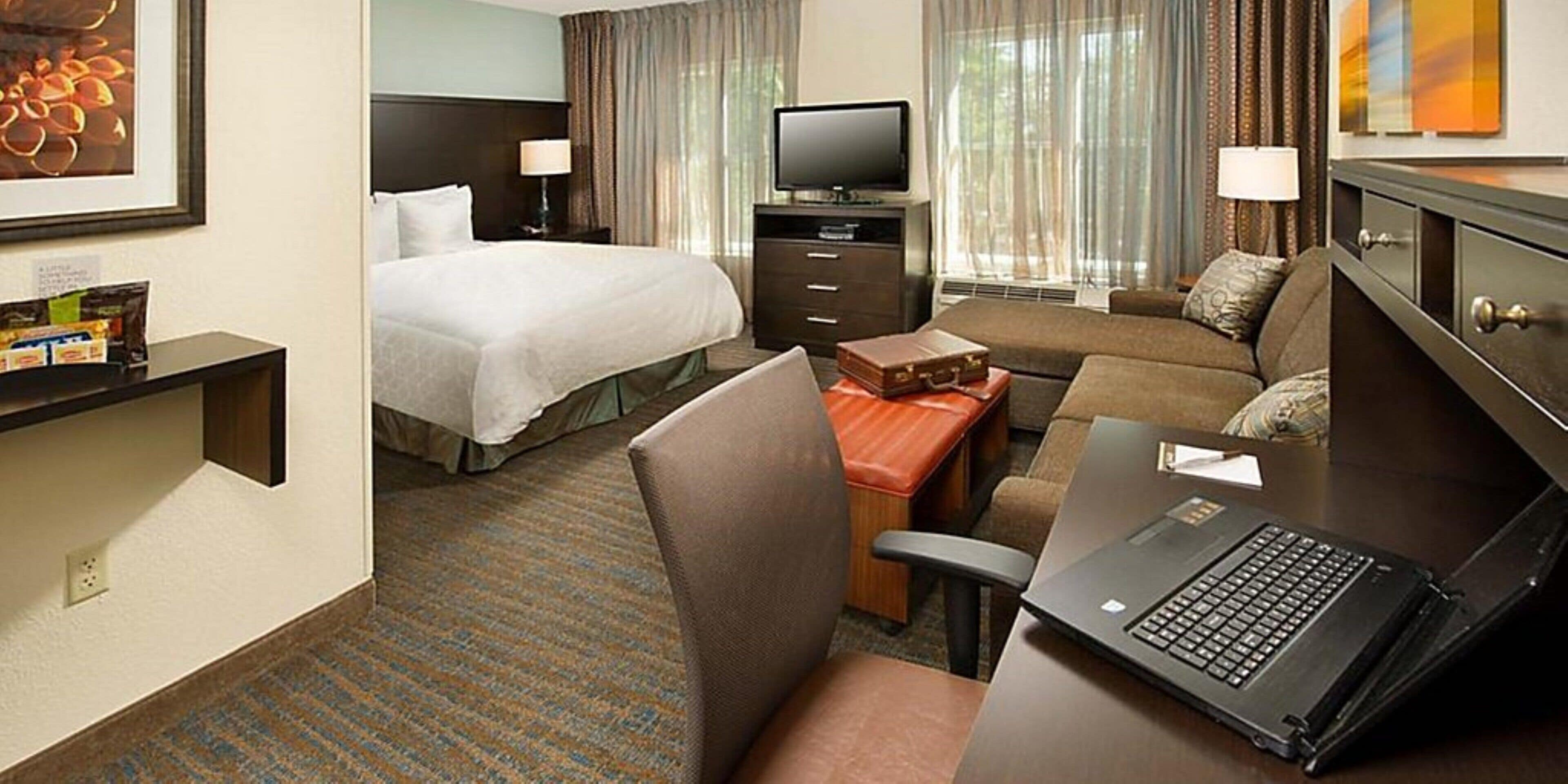 A hotel room with a bed, a desk with a laptop, a television, and a seating area with a couch and armchairs.