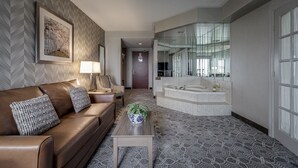 A modern hotel room with a brown leather sofa, patterned cushions, a small table with a vase, a large mirror, and a bathroom with a bathtub and a large mirror.