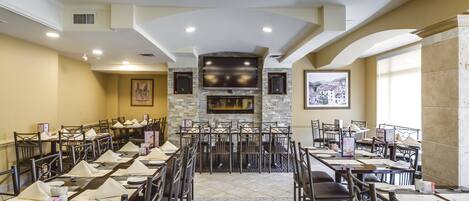 Un restaurante con una larga mesa de comedor, numerosas sillas, una chimenea de piedra y una pantalla de televisión grande.