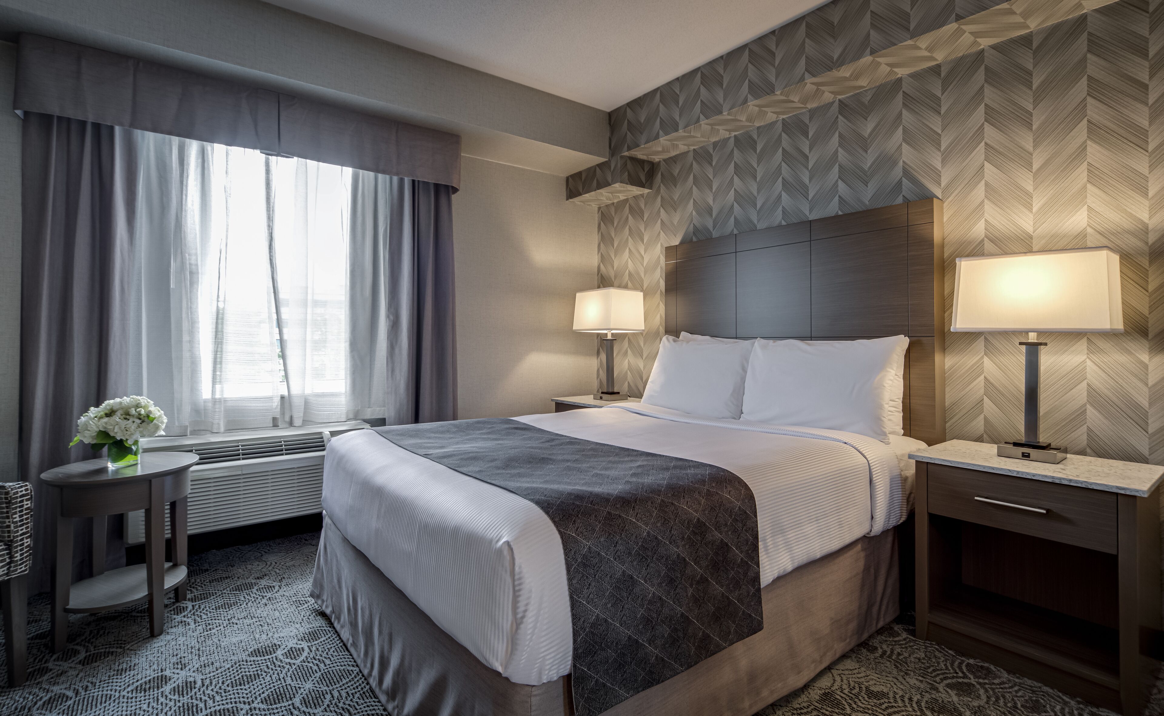 Habitación de hotel con una cama grande, dos lámparas de noche, una mesita con un florero y una pared con motivo de espiga.