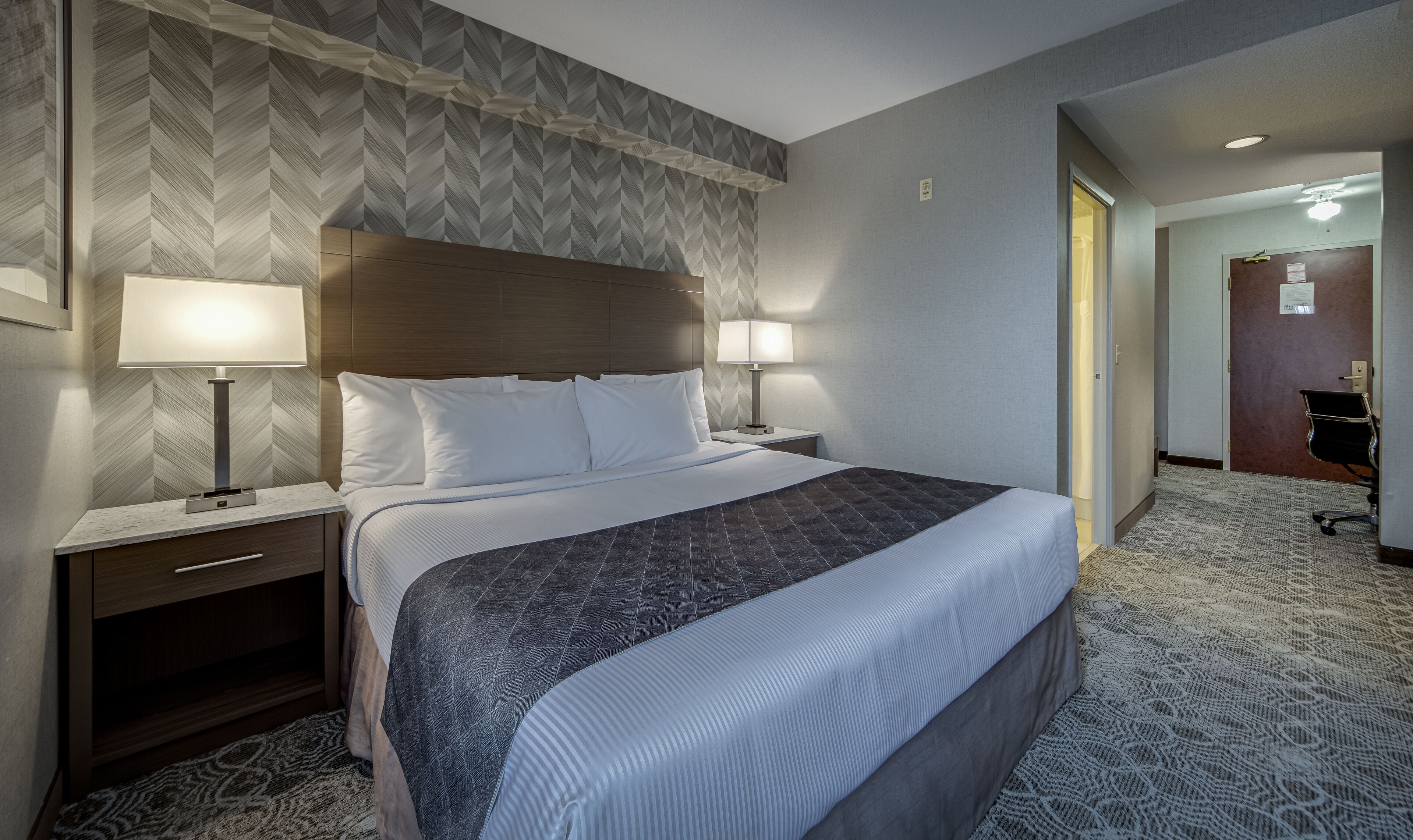 A hotel room with a large bed, two bedside lamps, a wooden headboard, and a patterned carpet.