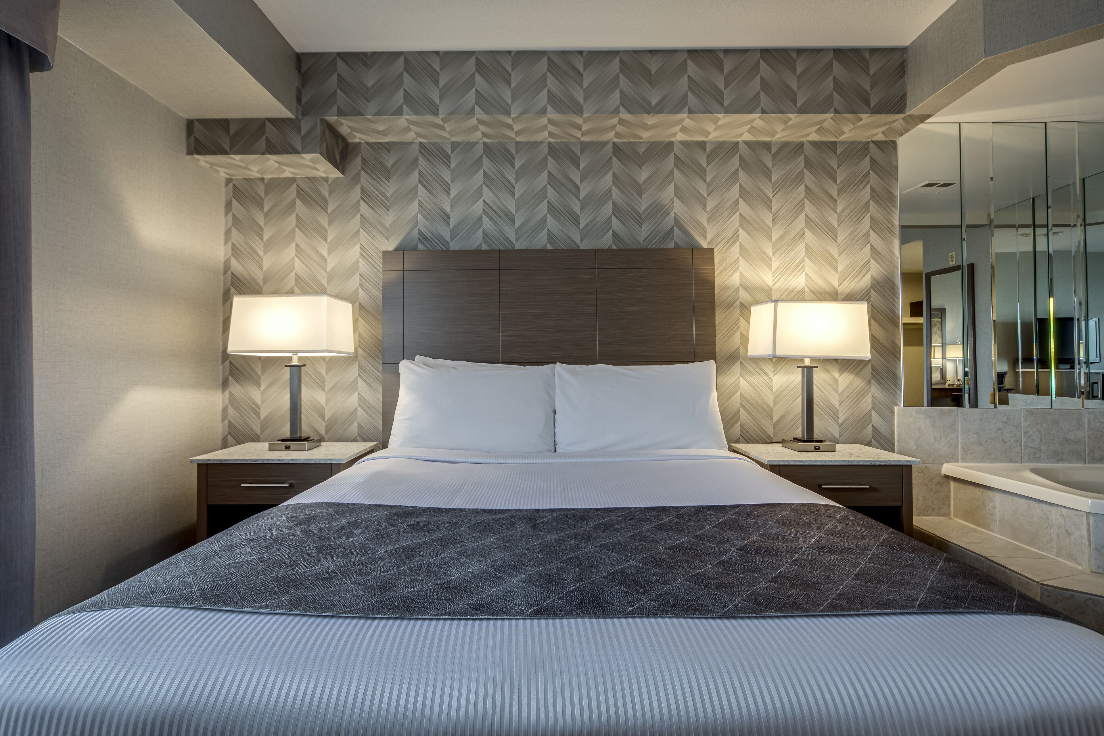 A hotel room with a large bed, two bedside tables with lamps, a modern headboard, and a large mirror above the bed.