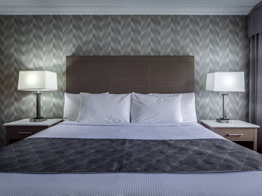 A hotel room with a large bed, two bedside tables with lamps, a wooden headboard, and a herringbone-patterned wallpaper.