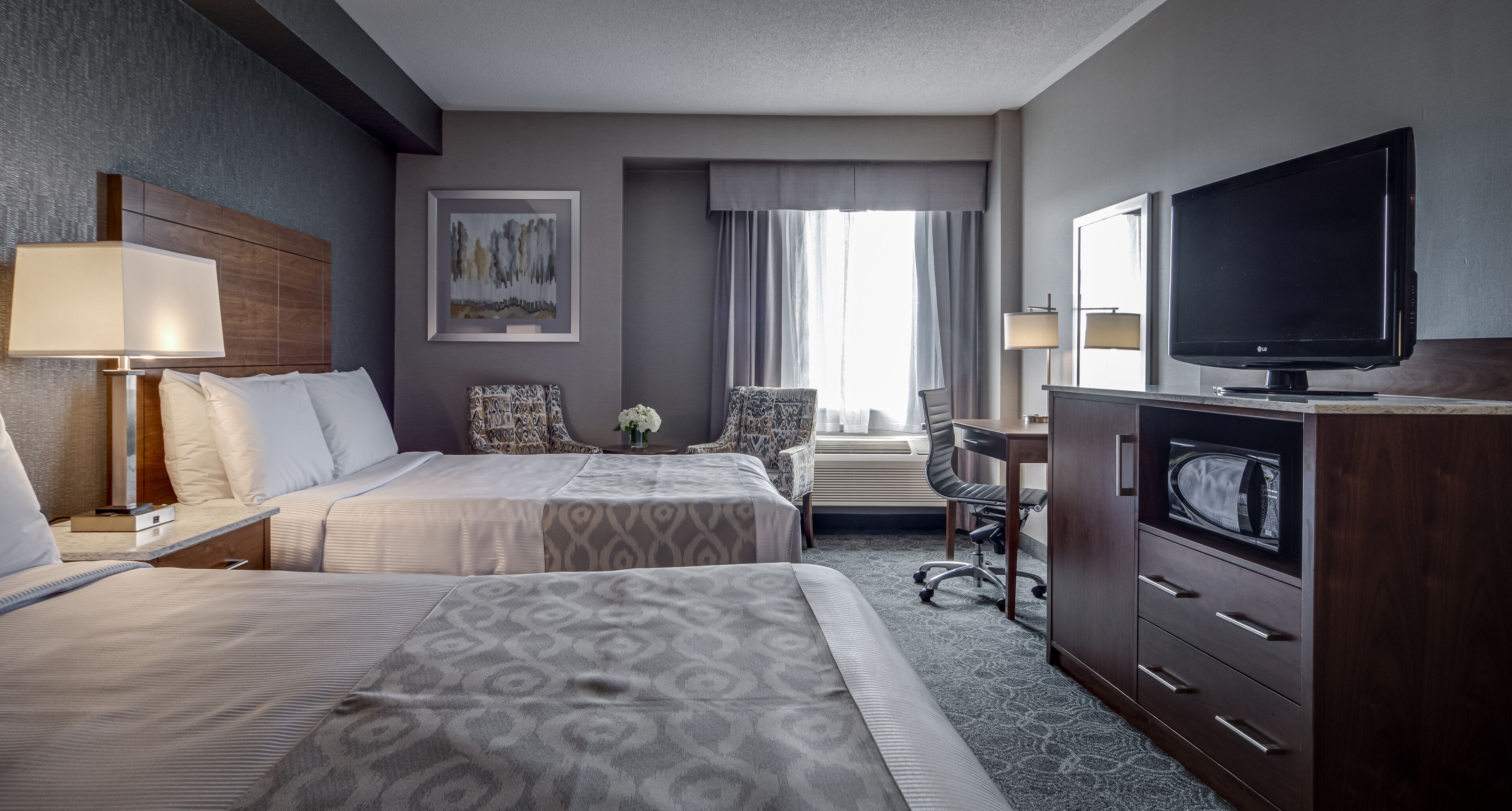 A hotel room with two beds, a desk, a television, and a large window with curtains.