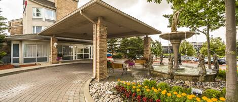 A hotel entrance with a covered patio, a fountain, and flower beds.