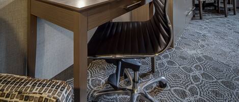 A hotel room with a wooden desk, a black office chair, a large mirror, a flat-screen TV, a framed picture on the wall, and a patterned carpet.