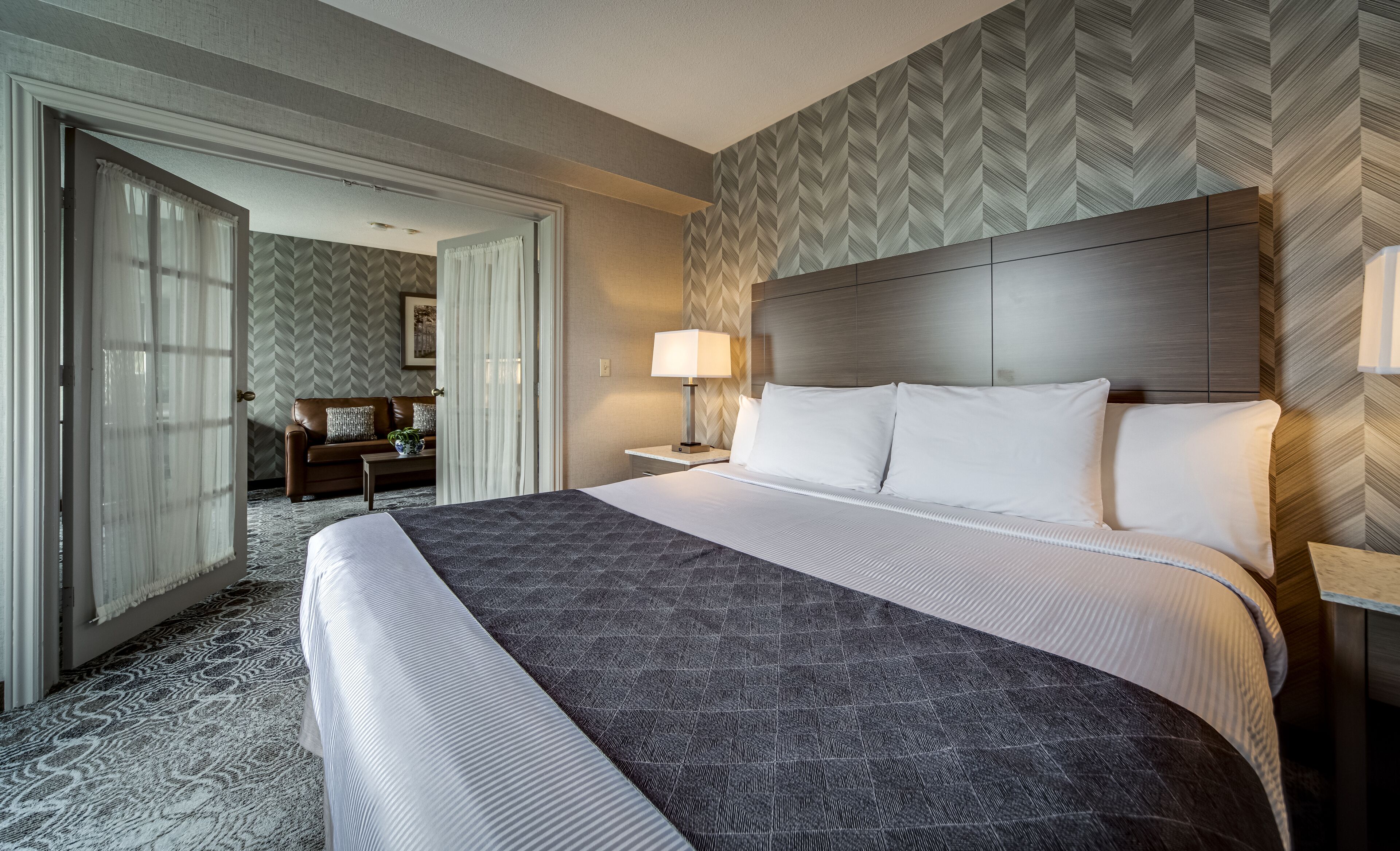 A hotel room with a large bed, a nightstand with a lamp, a sofa, and a herringbone-patterned wall.