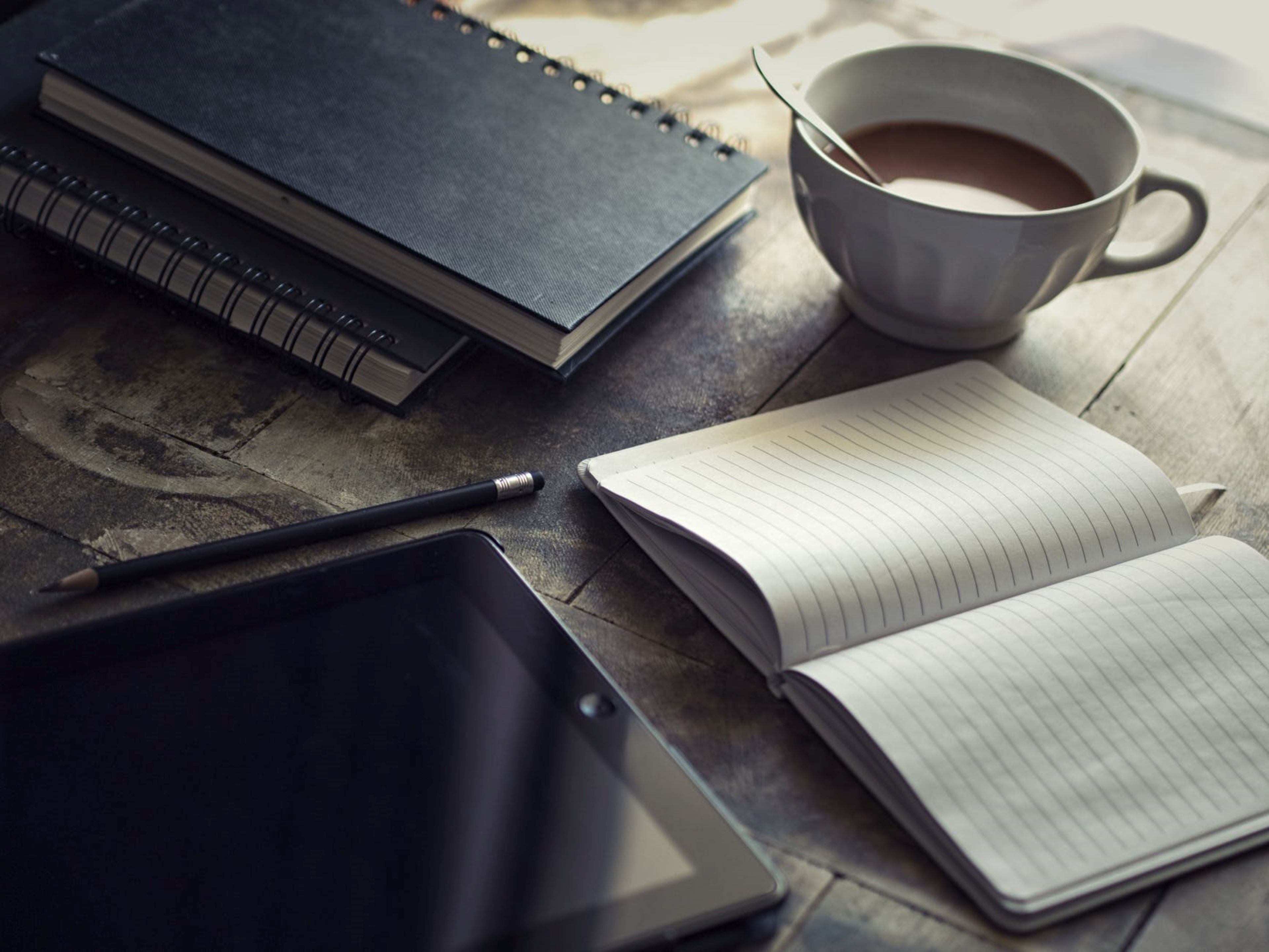 A notebook, a pen, a tablet, and a cup of coffee on a wooden table.