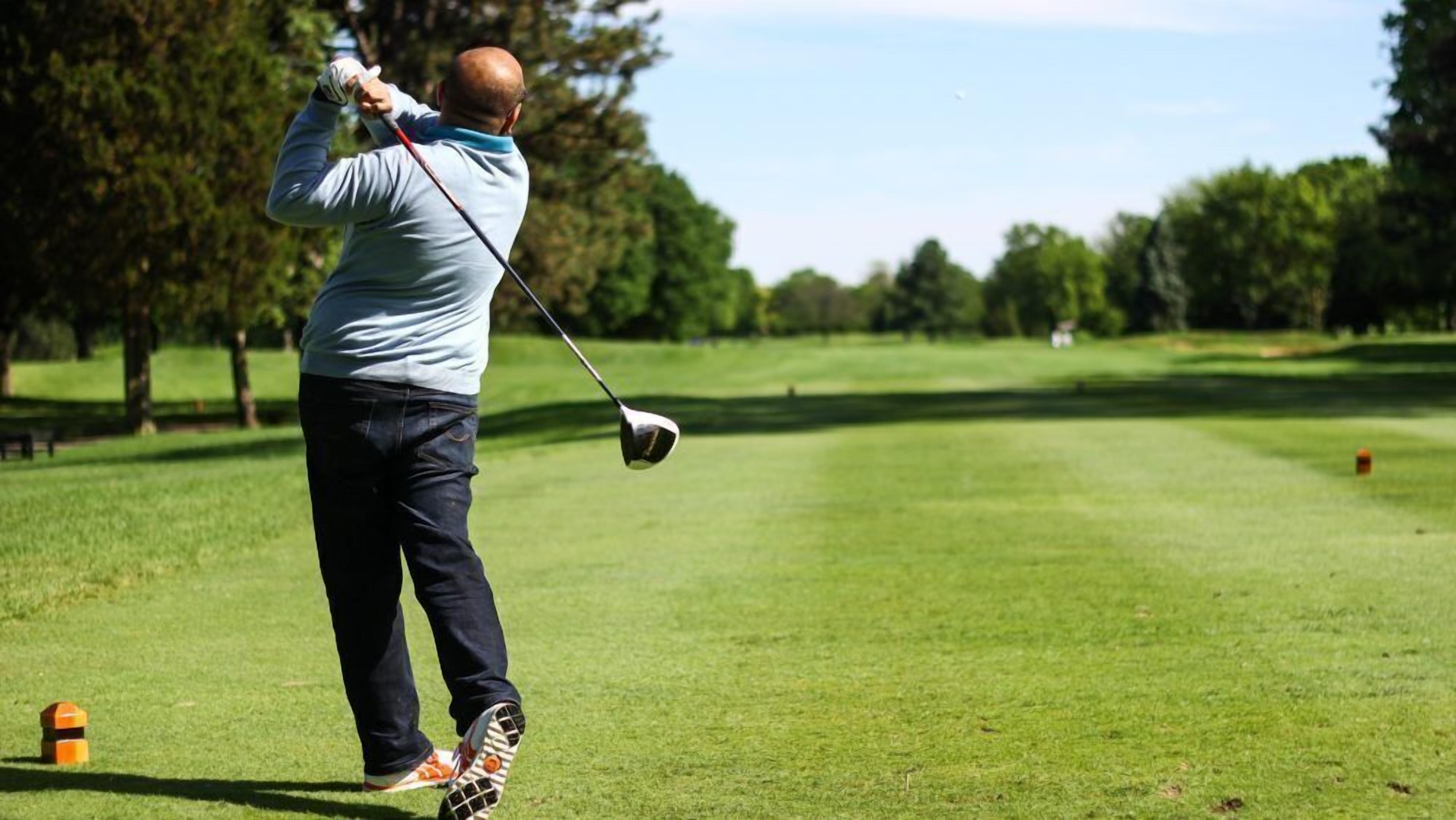 A golfer in mid-swing on a golf course with a clear sky and trees in the background.