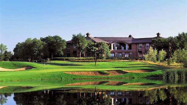 Un terrain de golf avec un étang dans lequel se reflète un bâtiment doté de nombreuses fenêtres et d’une façade en briques.