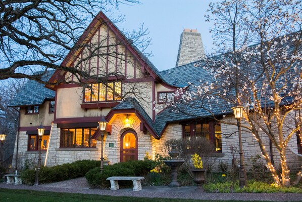 A two-story house with a stone chimney, a visible front door, and a well-maintained garden with a bench and a birdbath.