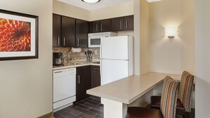 A compact kitchen with dark cabinets, a white refrigerator, a coffee maker, and a dining area with striped chairs. - Staybridge Suites Chicago - Glenview by IHG (Glenview)