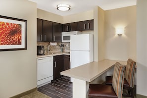 A compact kitchen with dark cabinets, a white refrigerator, a coffee maker, and a dining area with striped chairs. - Staybridge Suites Chicago - Glenview by IHG (Glenview)