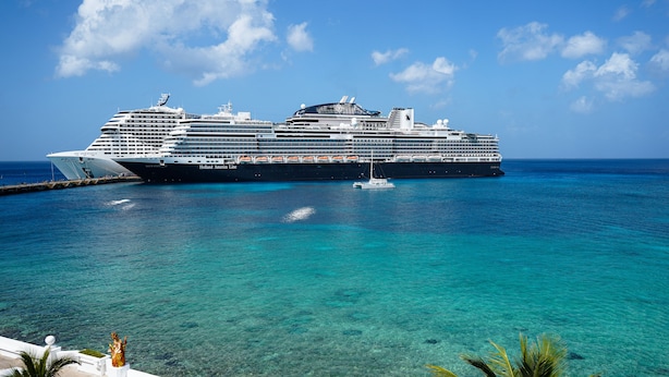 Un gran crucero amarrado en un puerto con aguas turquesas cristalinas y un edificio blanco con una estatua al frente.