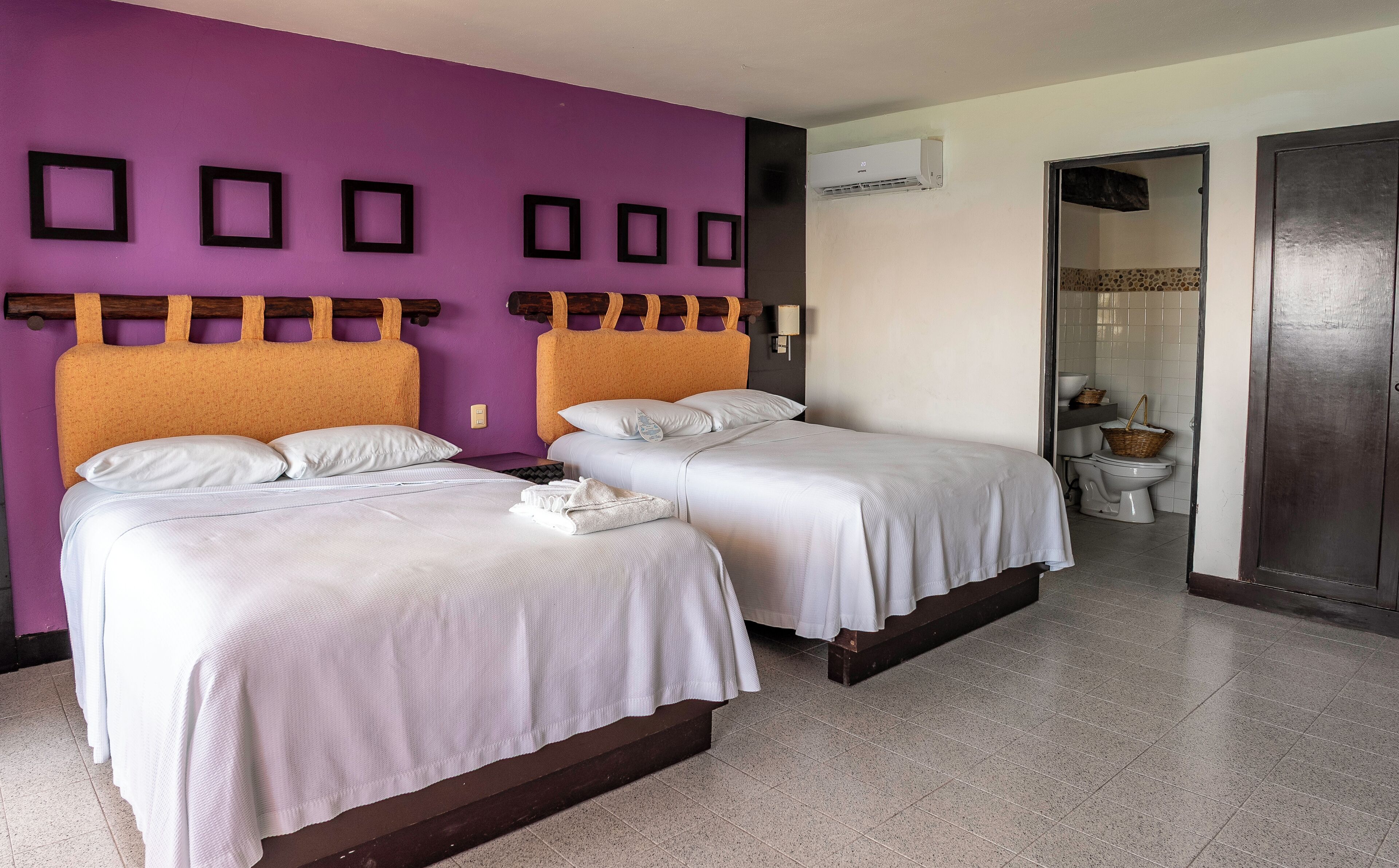 Two beds with white linens, a purple wall with black square decorations, and a bathroom visible through an open door.