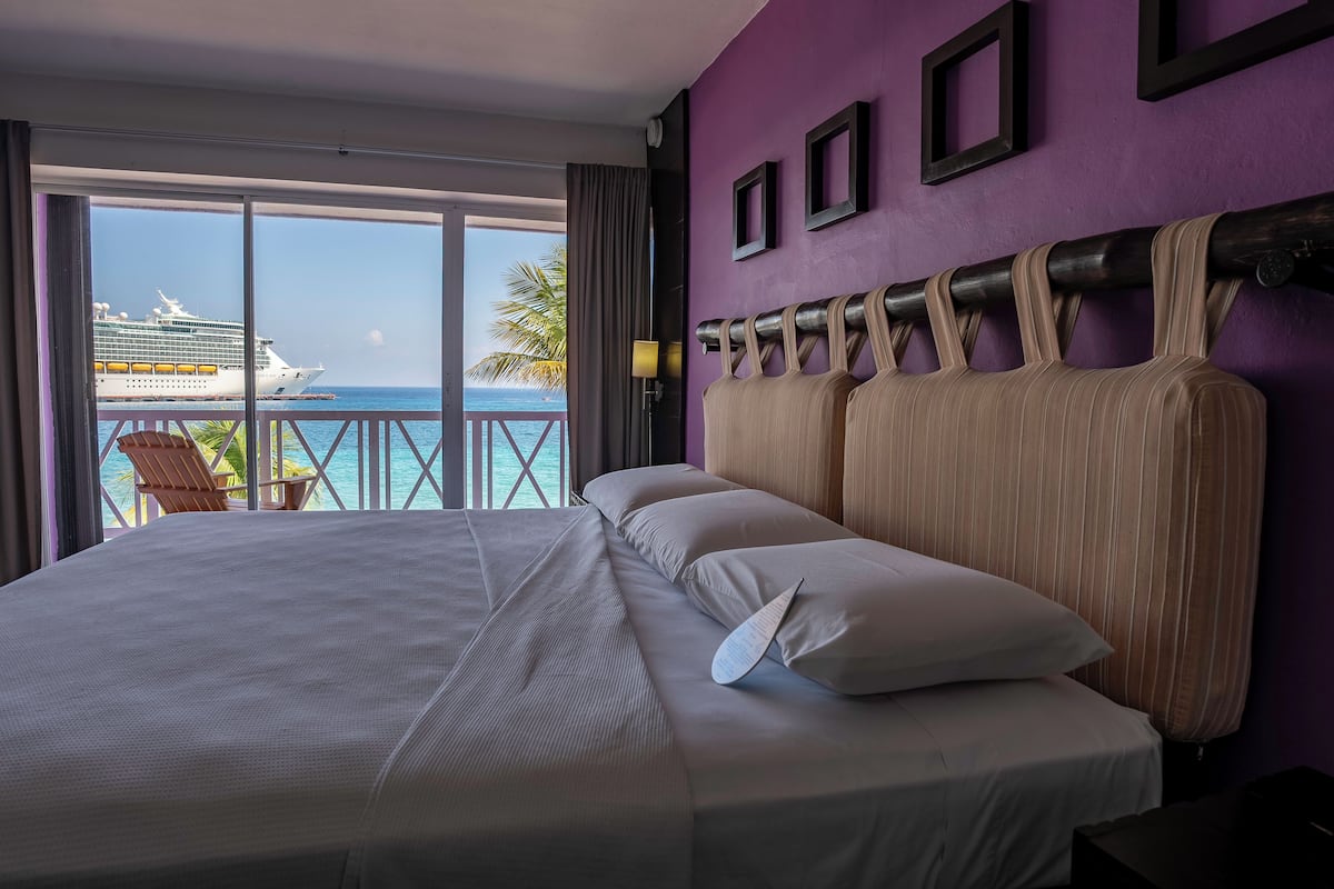 Een hotelkamer met een groot bed, uitzicht op de oceaan en een cruiseschip in de verte.