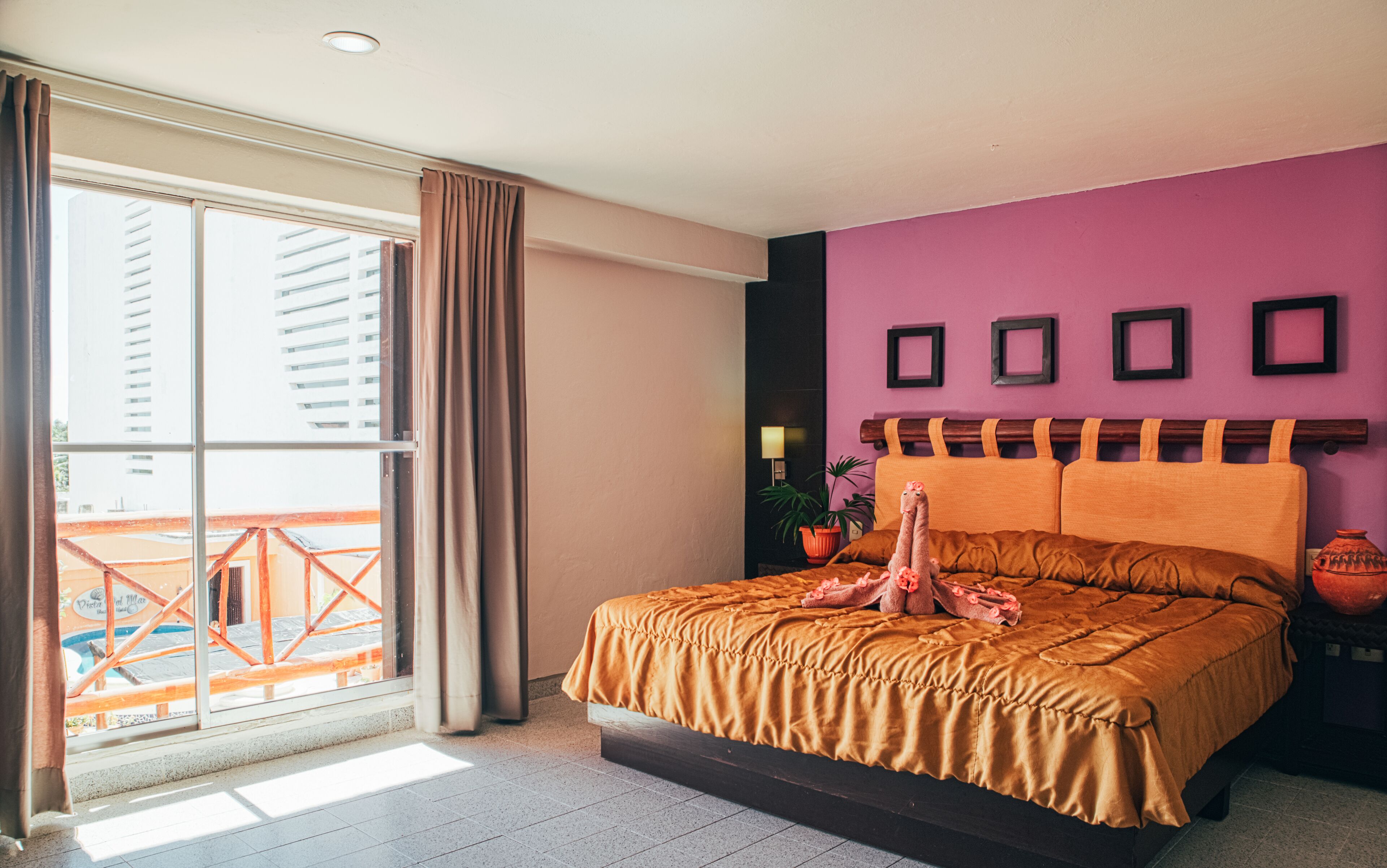 Een slaapkamer met een groot bed, een raam met gordijnen, uitzicht op gebouwen, en een paarse muur met ingelijste foto's.