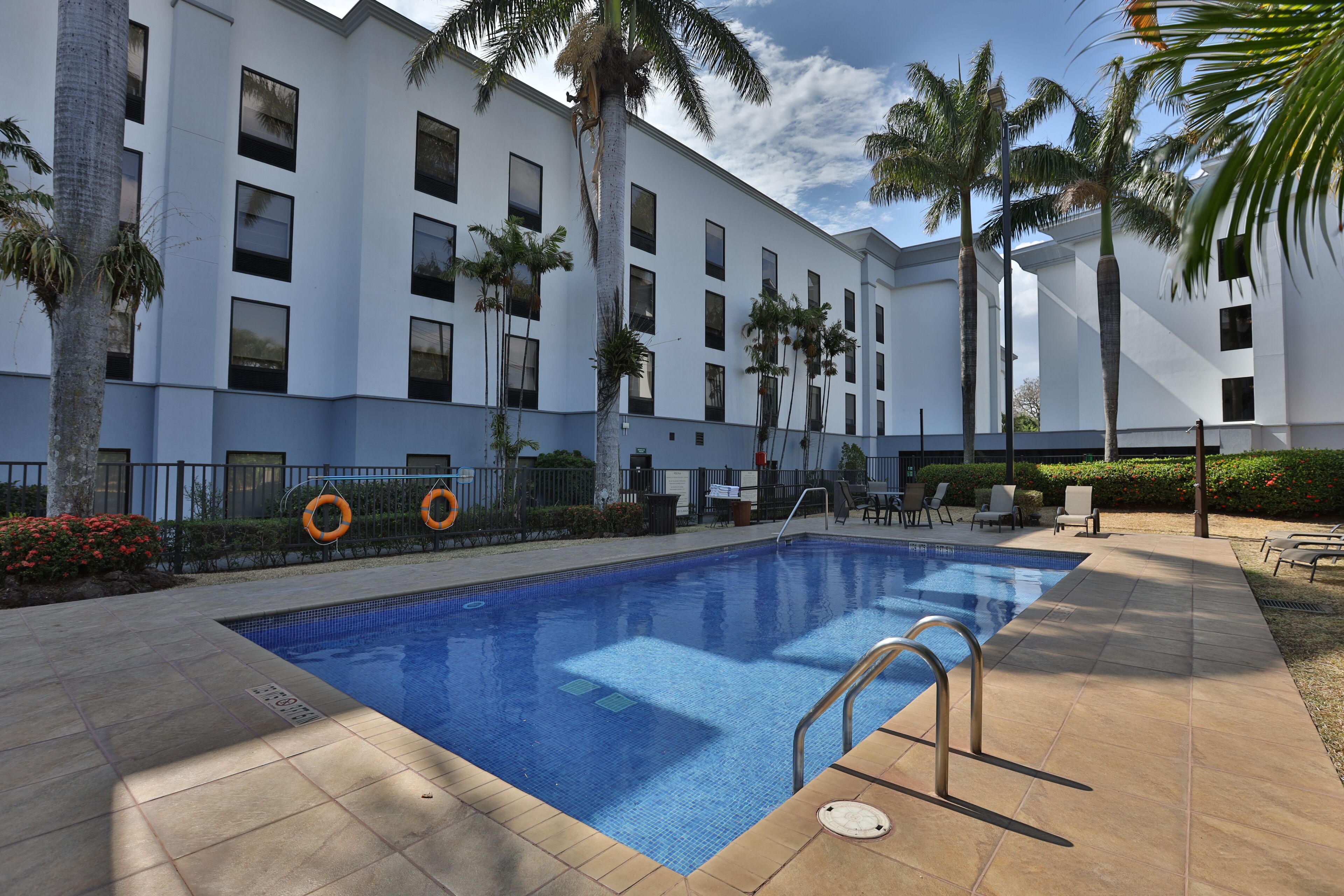 Una piscina con un bordeado de baldosas, palmeras y un edificio de fachada blanca con múltiples ventanas.