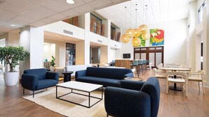Een ruime lobby met een modern design, met een zithoek met banken en een bijzettafeltje, een eetgedeelte met tafels en stoelen, en een grote plant in de hoek.