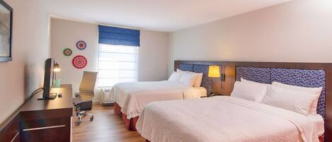 A hotel room with two beds, a desk, a chair, and a window with blinds.