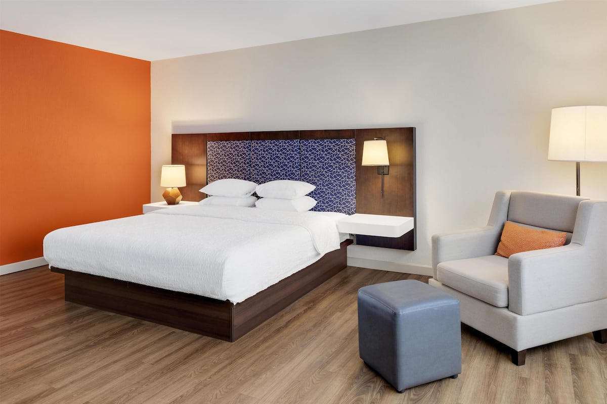 Ein Hotelzimmer mit einem großen Bett, einer Sitzecke mit einem Sessel, einem kleinen Hocker und einem Nachttisch mit Lampe.