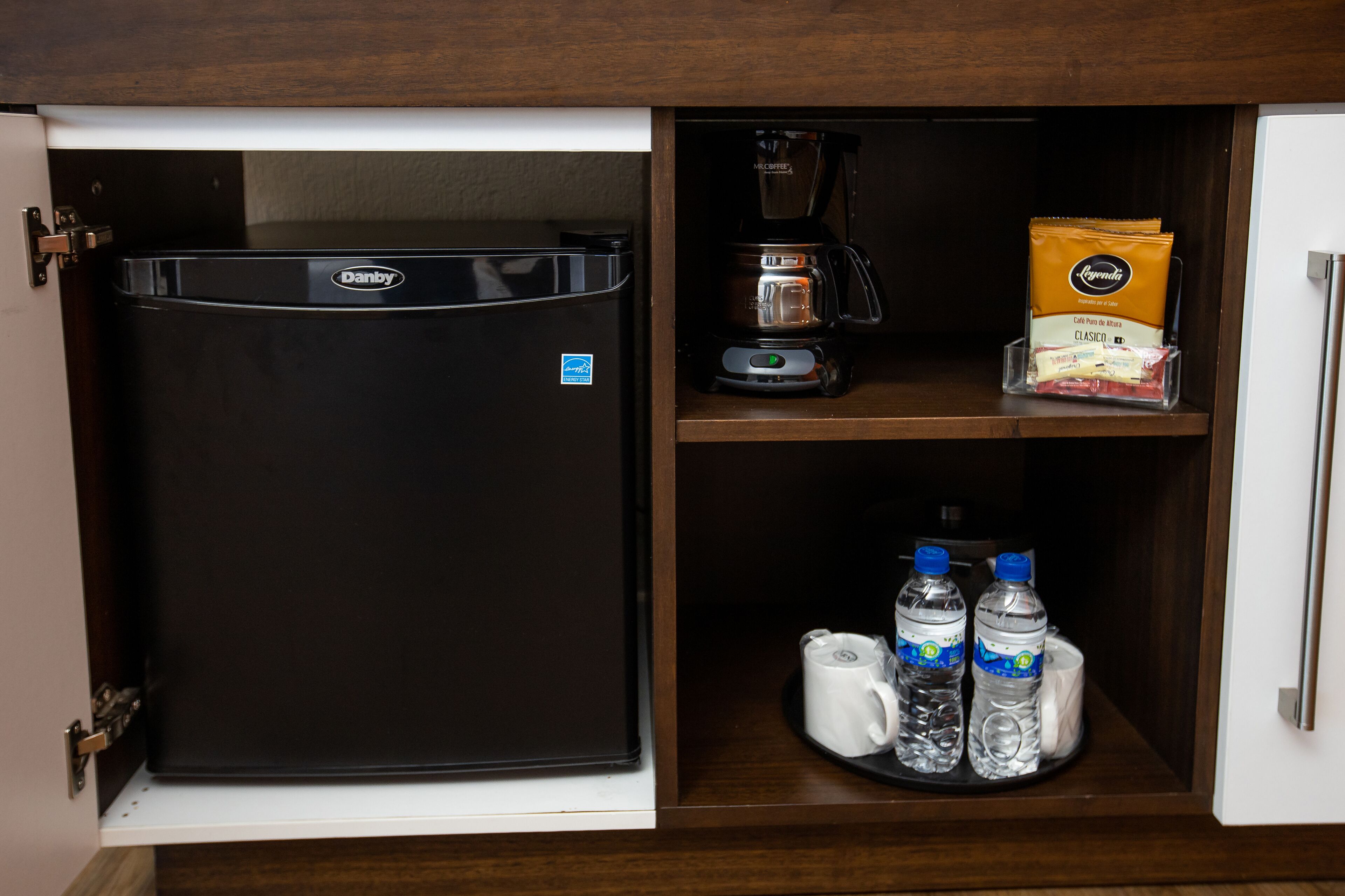 A compact kitchen with a black refrigerator, a coffee maker, a coffee pod dispenser, and bottled water.