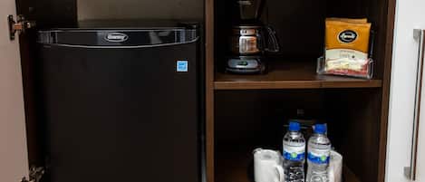 A compact kitchen with a black refrigerator, a coffee maker, a coffee pod dispenser, and bottled water.