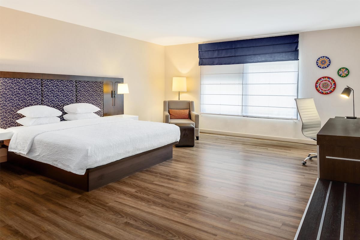 Ein Hotelzimmer mit einem großen Bett, einem Sessel, einem Schreibtisch und einem Fenster mit Vorhängen.