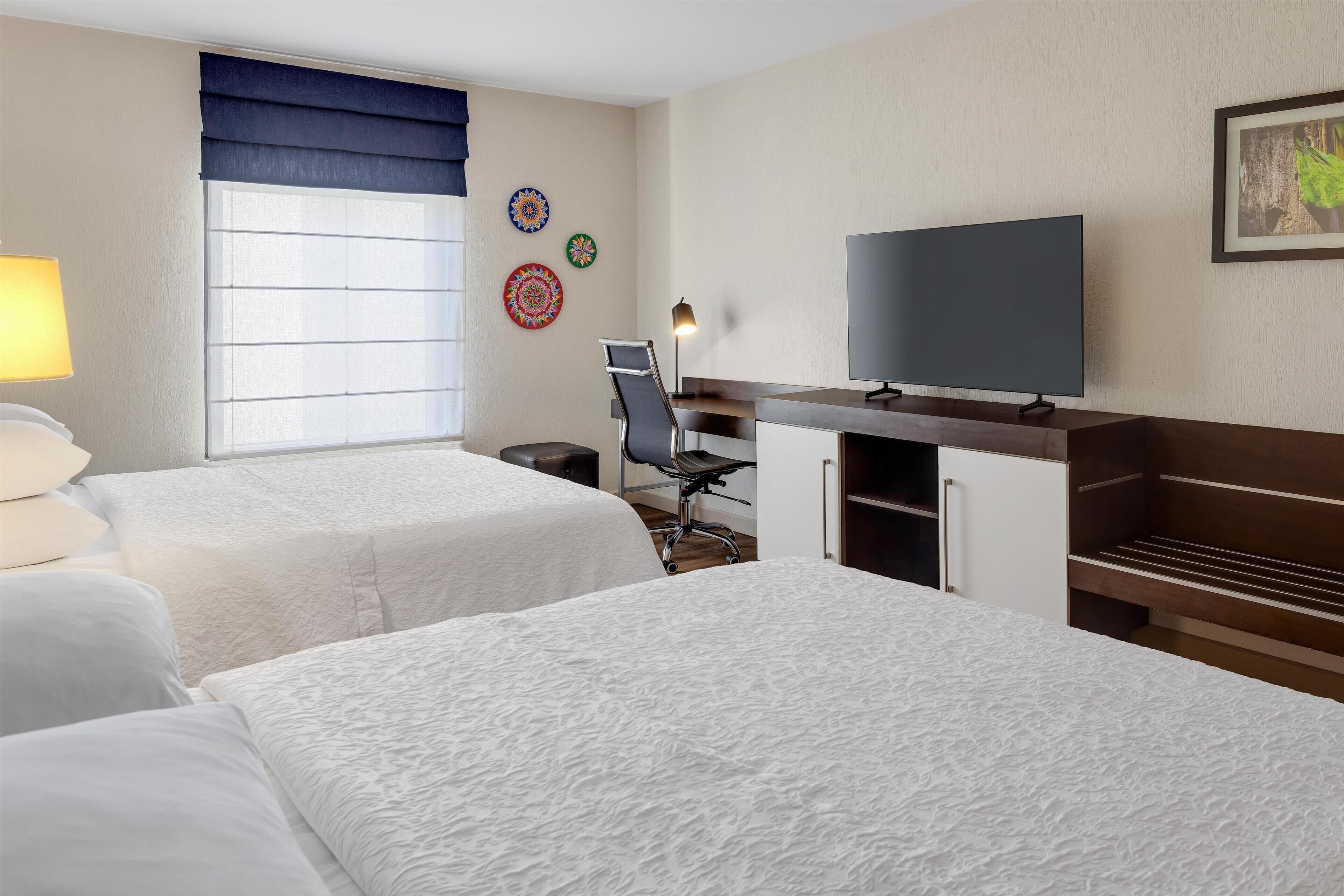 A hotel room with two beds, a desk with a computer, a TV, and a window with blinds.