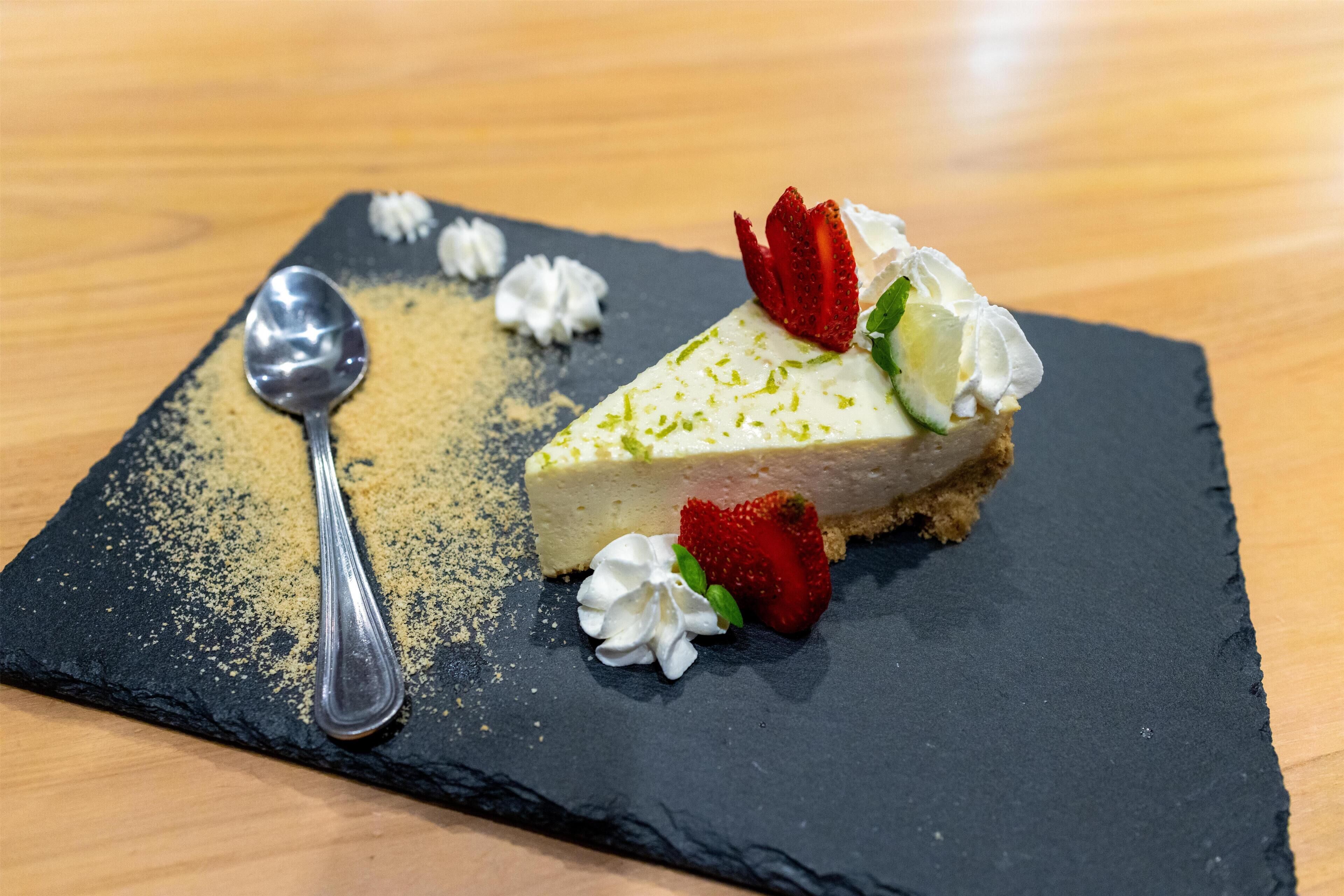 A slice of cheesecake on a slate plate with a spoon, garnished with whipped cream and strawberries.