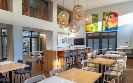 A modern restaurant with high ceilings, wooden tables and chairs, a central bar area, and artwork on the walls.