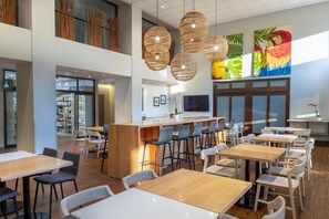 Un restaurante moderno con techos altos, mesas y sillas de madera, una zona de bar central y obras de arte en las paredes.