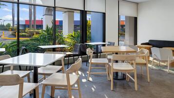Un área de estar interior moderna con mesas y sillas, ventanas amplias que ofrecen vistas al exterior, y una combinación de iluminación natural y artificial.