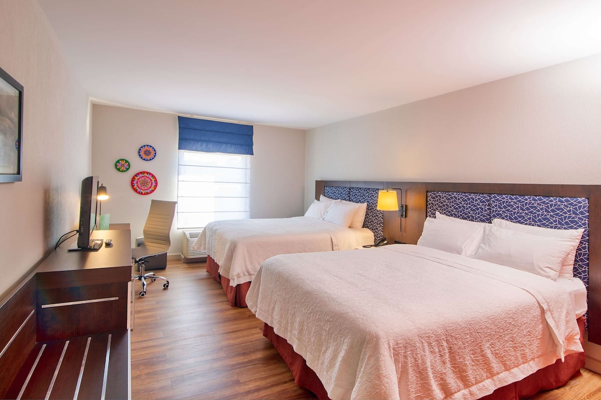 Ein Hotelzimmer mit zwei Betten, einem Schreibtisch, einem Stuhl, einem Fernseher und einem Fenster mit Jalousien.