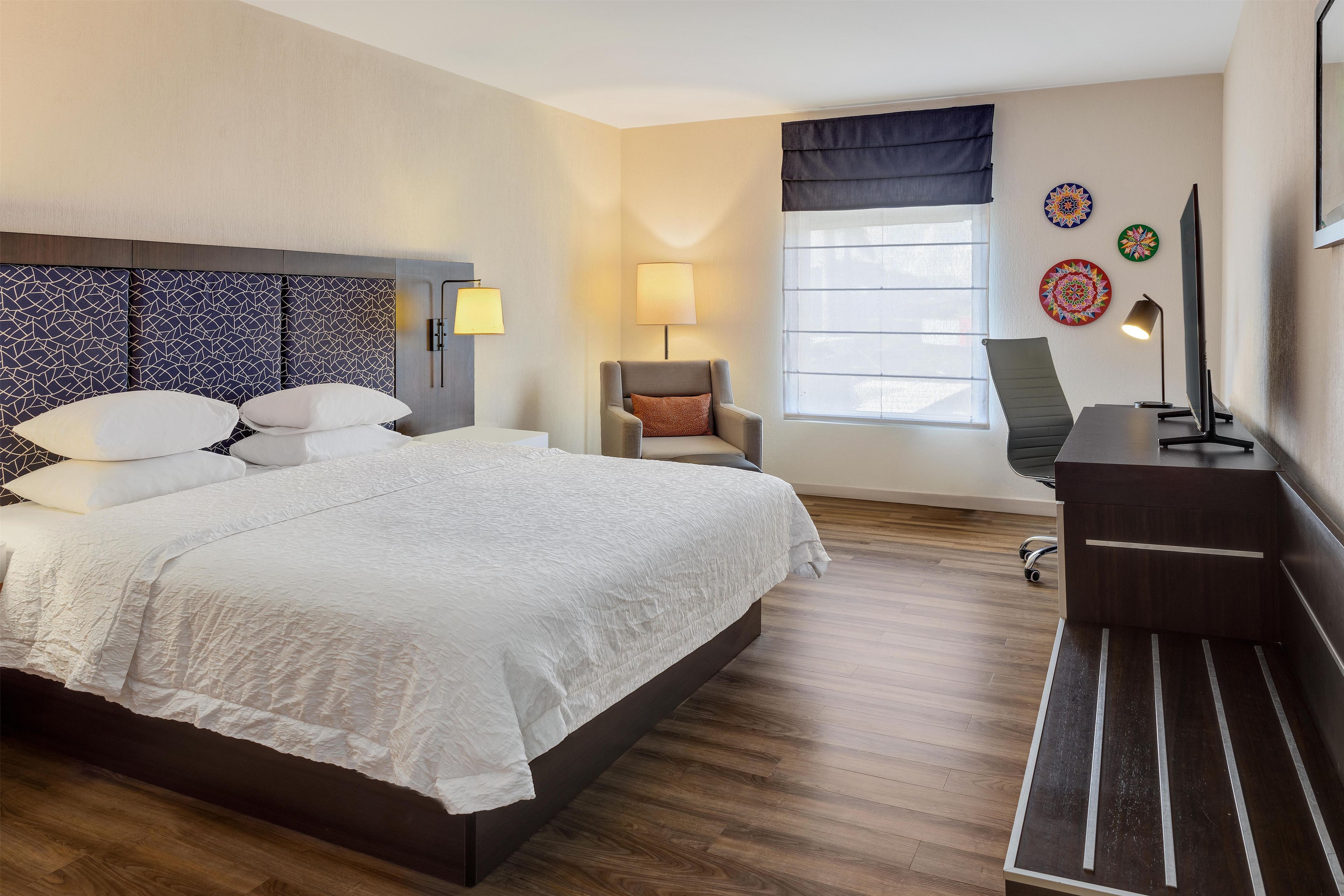 A hotel room with a large bed, a desk, a chair, and a window with blinds.